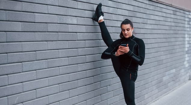 Young sportive girl with phone in black sportswear doing legs stretching outdoors by using gray wall.