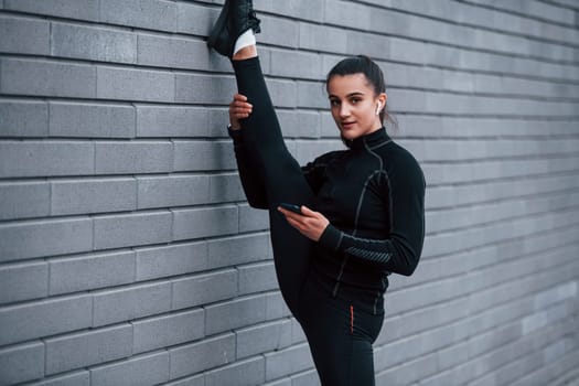 Young sportive girl with phone in black sportswear doing legs stretching outdoors by using gray wall.
