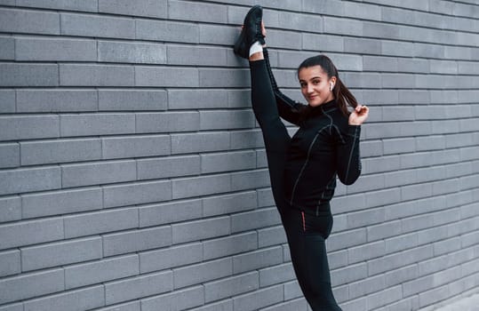 Young sportive girl in black sportswear doing legs stretching outdoors by using gray wall.