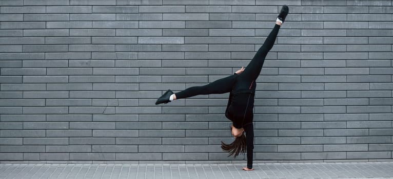 Young sportive girl in black sportswear doing hard handstand exercises outdoors near gray wall.