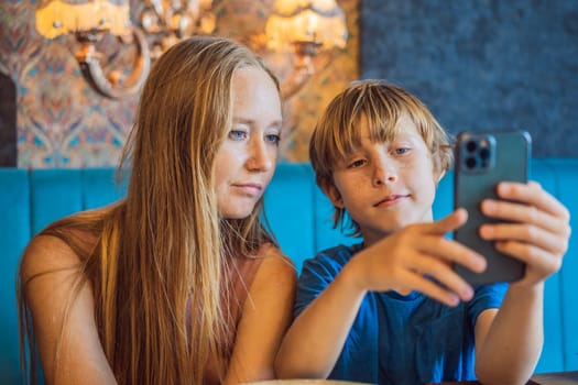 Caring young mum sit on couch in living room with small son watch funny cartoon on smartphone, happy mother or nanny relax on sofa at home with little boy child use cell browse Internet together.