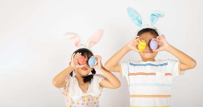 Happy Easter Day. Two smile Asian little girl and boy wearing easter bunny ears holding colorful eggs closes eyes with testicles isolated on white background with copy space, Happy child in holiday