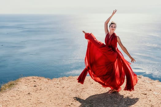 Side view a Young beautiful sensual woman in a red long dress posing on a rock high above the sea during sunrise. Girl on the nature on blue sky background. Fashion photo.