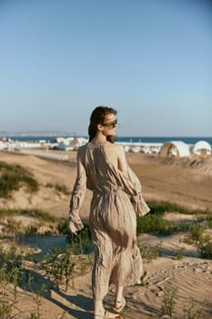 an elegant woman in a beige dress walks on the sand against the backdrop of the sea visible in the distance. High quality photo