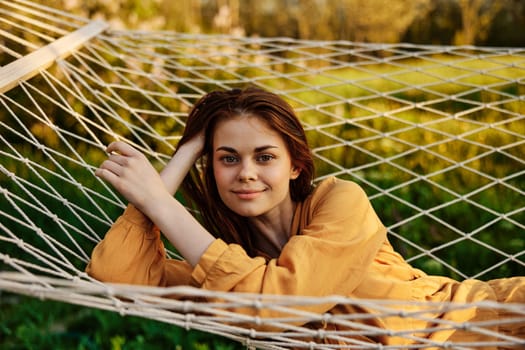 a happy woman is resting in a mesh hammock, resting her head on her hand, smiling happily looking at the camera, enjoying a sunny day. High quality photo
