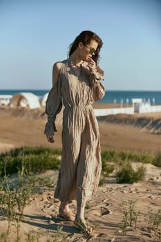 a sophisticated woman in a light beige dress flying in the wind is standing on the sand near the sea in sunglasses. High quality photo