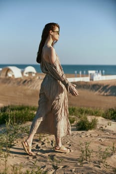 a sophisticated woman in a light beige dress flying in the wind is standing on the sand near the sea in sunglasses. High quality photo