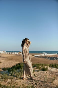 a sophisticated woman in a light beige dress flying in the wind is standing on the sand near the sea in sunglasses. High quality photo