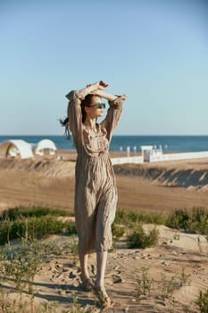 a slender, sophisticated woman in a beige dress stands on the sand in windy weather with her hands raised to her head. High quality photo
