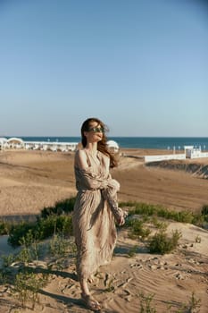 an elegant woman in a beige dress walks on the sand against the backdrop of the sea visible in the distance. High quality photo