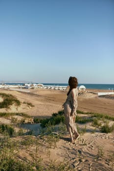 an elegant woman in a beige dress walks on the sand against the backdrop of the sea visible in the distance. High quality photo