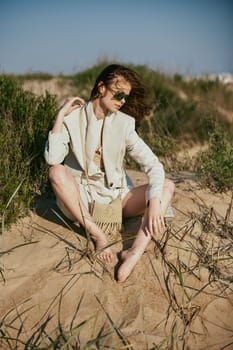 a beautiful woman in a light, fashionable jacket, in sunglasses sits barefoot on the sand in windy weather. High quality photo