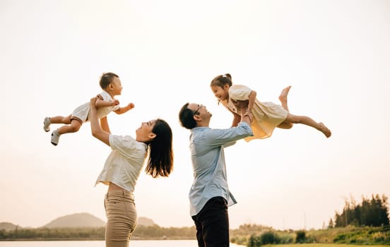 A cheerful Asian family enjoys a day out in nature, as the father and mother throw their toddler up into the sunny sky. The happy child enjoys the playful freedom of flying, captured in a photograph