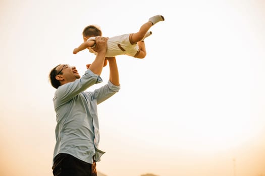 On a spring day in nature, a loving father holds his little girl up high in the park. The toddler daughter throws her arms up in playful freedom, enjoying a moment of love and happiness with her dad