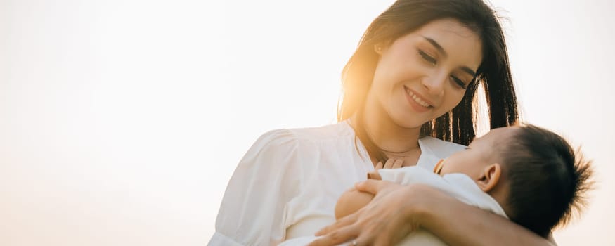 A heartwarming Mother's Day moment captured in the park at sunset. Beautiful mother cradles her sleeping baby, basking in peaceful nature and lovingly smiling at her newborn. Family care at its finest