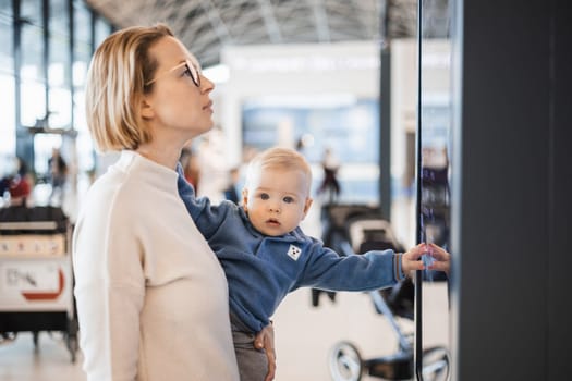 Mother traveling with child, holding his infant baby boy at airport terminal, checking flight schedule, waiting to board a plane. Travel with kids concept