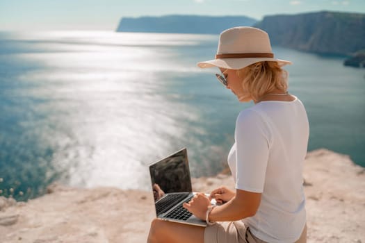 Freelance women sea working on the computer. Good looking middle aged woman typing on a laptop keyboard outdoors with a beautiful sea view. The concept of remote work