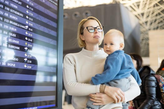 Mother traveling with child, holding his infant baby boy at airport terminal, checking flight schedule, waiting to board a plane. Travel with kids concept