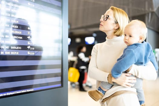 Mother traveling with child, holding his infant baby boy at airport terminal, checking flight schedule, waiting to board a plane. Travel with kids concept