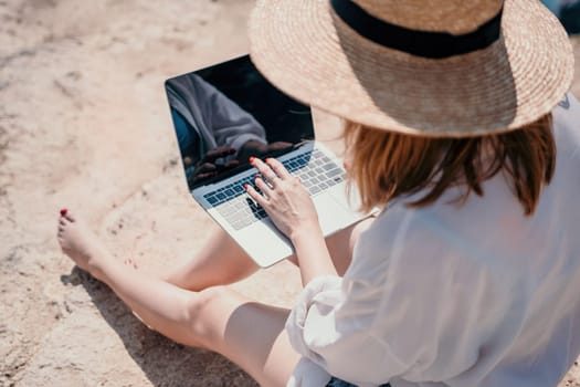 Successful business woman in yellow hat working on laptop by the sea. Pretty lady typing on computer at summer day outdoors. Freelance, travel and holidays concept.