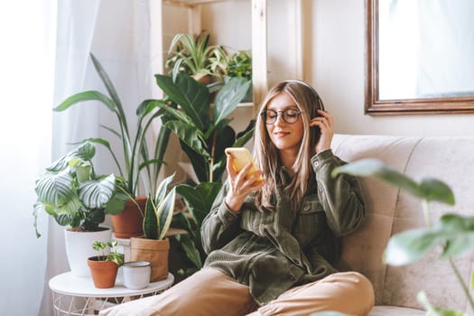 Freelance woman in glasses with mobile phone listening music in headphones and relax at home. Happy girl with closed eye sitting on couch in living room with plants.
