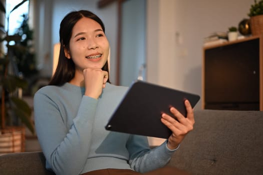 Happy asian female freelancer holding digital tablet looking outside through window with joyful smile.