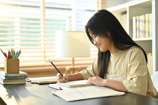 Image of smiling asian teenage student, doing homework, writing task, taking notes on desk at home.