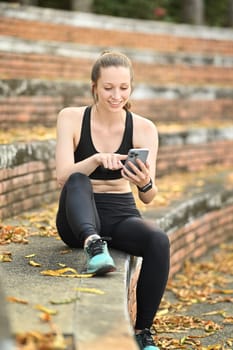 Beautiful caucasian female runner in sport clothes using smart phone, resting after morning workout.