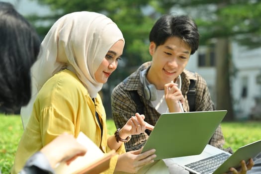 Smiling Muslim student and her friend using digital tablet on green meadow in campus. Education, youth and community concept.