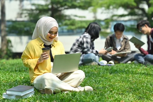 Happy young muslim student using laptop on green meadow in campus. Education technology and community concept.