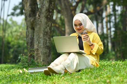 Full length of young asian muslim woman using laptop at summer park, browsing social networks and working online.