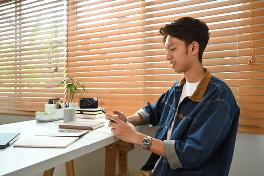 Young hipster man sitting in cozy home office interior and using smart phone. Freelance, technology and lifestyle concept.