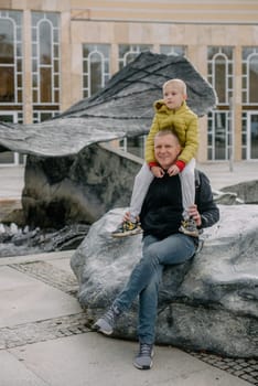 Dad holds on his son's shoulders. Beautiful family is spending time together outside. Dad and his little son are having fun on a roof terrace with view on a city. Sitting on father's shoulders and smiling.