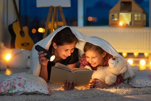 Happy mother with little daughter reading book using flashlight under blanket on floor in children's room