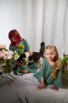 Amazing pretty mother and daughter having fun with flowers and cat in 8 March or in Mother's day. Red haired mom and small little blonde girl having lovely time on white background in studio with pet