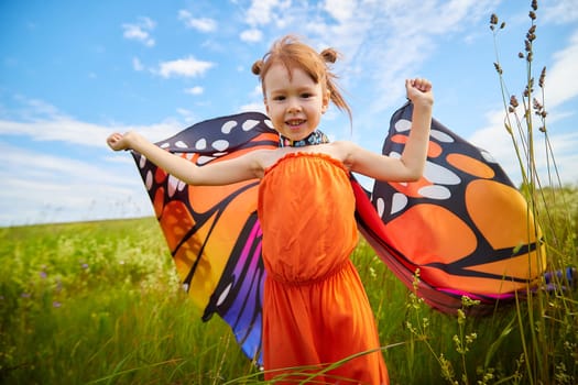 Portrait of little girl with Asian eyes and butterfly wings having fun and joy in meadow or field with grass, flowers on sunny summer day