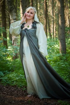 Adult mature woman 40-60 in a green long fairy dress in forest. Photo shoot in style of dryad and queen of nature. Fairy in beautiful green summer forest. Concept of caring for nature