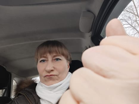 Portrait of adult cute funny blonde woman in warm clothes taking a selfie inside the car and driving it. The concept of pleasant trip in spring, autumn, winter