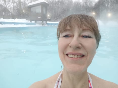 Adult mature woman takes selfie in a pool with warm hot termal mineral water in winter and white snow around. Wellness center and the concept of health care. Travel, recreation, medicine and rest