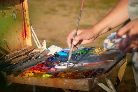 Box easel with canvas, oil paint, palette and tools and Hand of painter with brush on nature outdoors