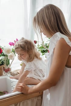 A little blonde girl with her mom on a kitchen countertop decorated with peonies. The concept of the relationship between mother and daughter. Spring atmosphere