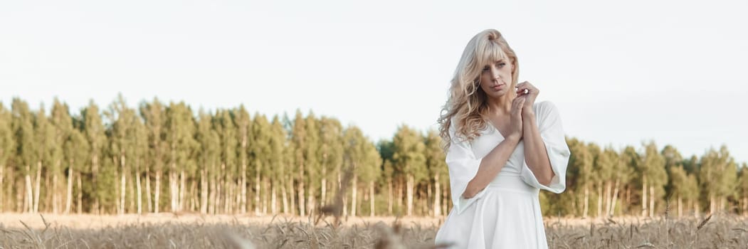 A blonde woman in a long white dress walks in a wheat field. The concept of a wedding and walking in nature.