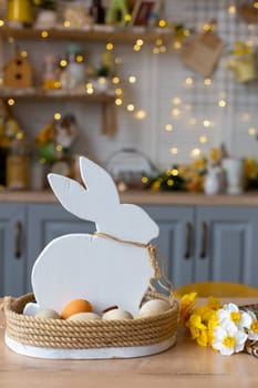A basket with a wooden white rabbit and natural eggs stands on the table in the kitchen, with Easter decorations and flowers. Vertical. Close up
