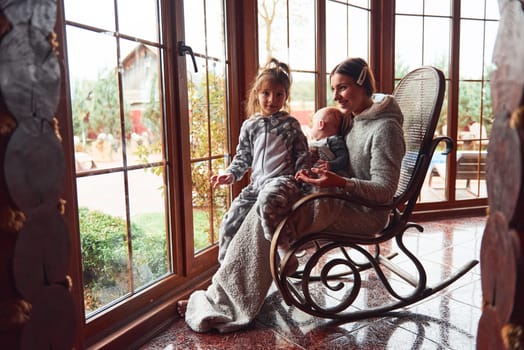 Happy mother sits with her little son and daughter on the chair near window.