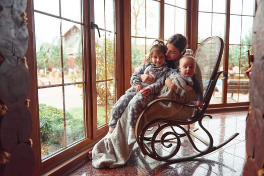 Happy mother sits with her little son and daughter on the chair near window.