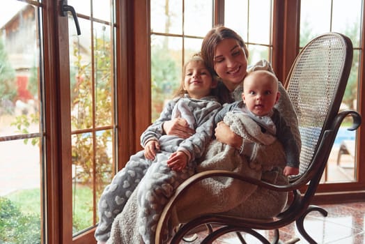 Happy mother sits with her little son and daughter on the chair near window.