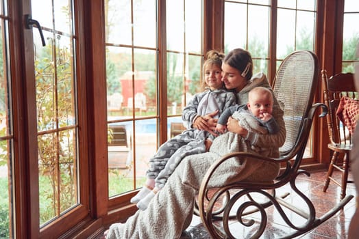 Happy mother sits with her little son and daughter on the chair near window.