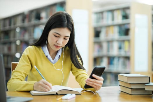 Young female college highschool student with headphones and in casual cloths sitting at desk studying, reading books, and writing with handphone at library for research or school project. E-Learning Education Library concept.