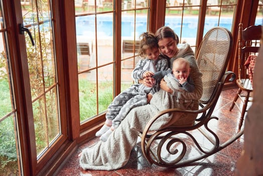 Happy mother sits with her little son and daughter on the chair near window.