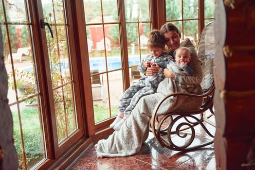 Happy mother sits with her little son and daughter on the chair near window.